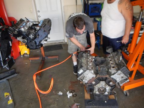 2003 Ford Crown Victoria - man crouches between two V8 engine blocks on a garage floor, working with tools and an orange air hose.