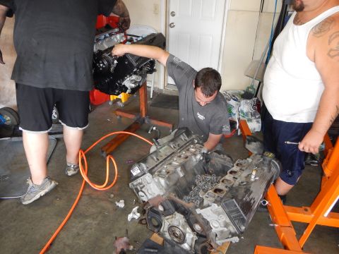 2003 Ford Crown Victoria - Men work between two V8 engines, one mounted on an orange stand and one on the garage floor, surrounded by tools.