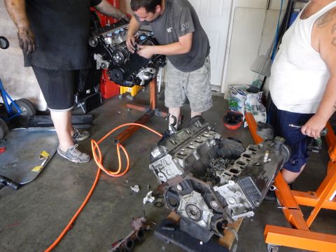 2003 Ford Crown Victoria - man bends over a V8 engine on an orange stand while a second V8 block sits on the garage floor amid scattered tools.