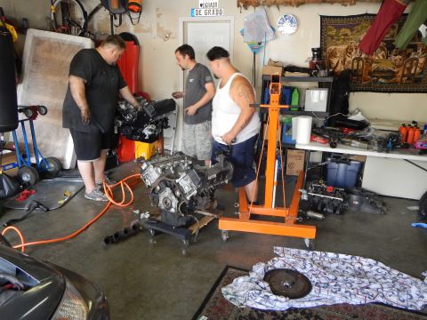 2003 Ford Crown Victoria - Three men gather around a V8 engine on an orange hydraulic engine stand in a cluttered garage with a tapestry on the wall.