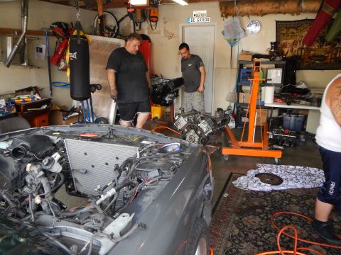2003 Ford Crown Victoria - Two men stand in a cluttered garage next to a gray sedan with its front end stripped, with a V8 engine and orange hoist behind them.