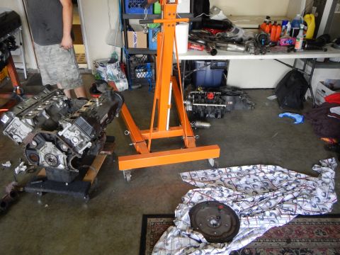 2003 Ford Crown Victoria - orange engine hoist or stand sits between two V8 engines resting on the garage floor near a flywheel on a sheet.