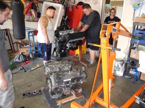 2003 Ford Crown Victoria - Several people work on two V8 engines in a garage, one mounted upright on an orange engine stand and one on the floor.
