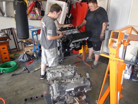 2003 Ford Crown Victoria - Two men work on a V8 engine mounted on an orange stand while a second V8 engine sits on the garage floor below.