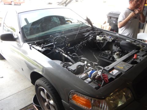 2003 Ford Crown Victoria - gray sedan sits with its hood raised and engine removed, showing the bare engine bay and wiring as a man stands at right.