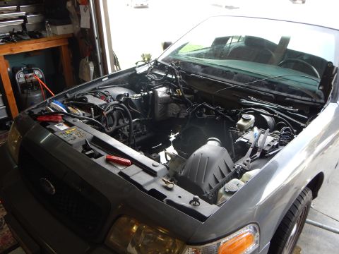 2003 Ford Crown Victoria - gray sedan with hood raised and engine pulled, showing the empty engine bay; a Ford oval badge is visible on the grille.