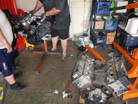 2003 Ford Crown Victoria - man hoists a V8 engine on an orange stand while a second disassembled V8 with timing chains lies on the garage floor.