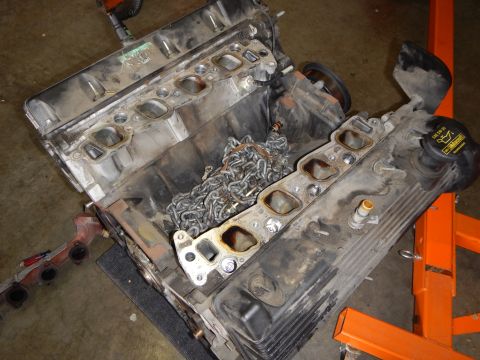 2003 Ford Crown Victoria - grimy V8 engine block sits with cylinder heads removed, exposing intake ports and timing chains beside an orange stand.