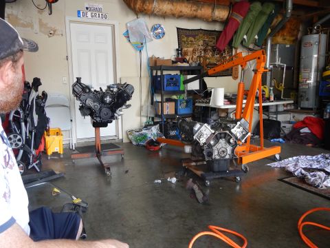 2003 Ford Crown Victoria - Two V8 engines sit on stands in a garage with an orange engine hoist between them as a man in a cap watches from the left.
