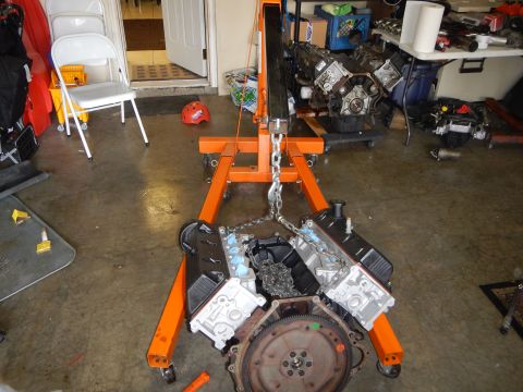 2003 Ford Crown Victoria - orange engine hoist lifts a V8 engine by a chain, the flywheel facing the camera, with more engines on a bench behind.