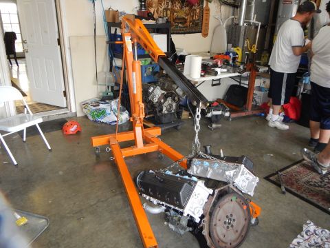 2003 Ford Crown Victoria - orange engine hoist suspends a V8 engine by a chain, flywheel visible, while several people work in the garage behind.