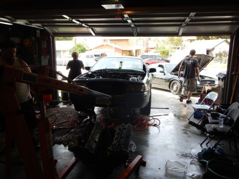 2003 Ford Crown Victoria - backlit open garage shows a stripped dark sedan with hood up, an engine on a stand, and cars parked in the driveway beyond.