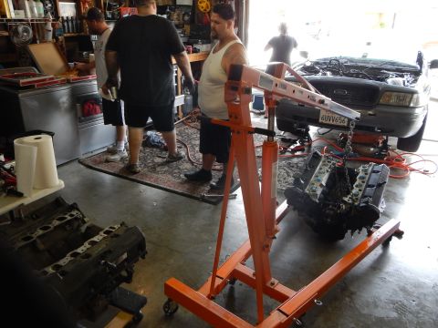 2003 Ford Crown Victoria - Men work in a garage with an orange hoist and V8 engine; a dark sedan with plate 4UVV656 sits with its hood up behind them.