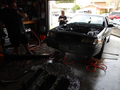2003 Ford Crown Victoria - black Ford sedan with California plate sits hood-up in a home garage as two people work on its partly disassembled engine.