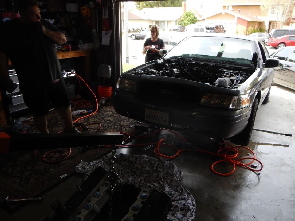 Black Ford sedan in a garage with hood up, engine partly disassembled, people working