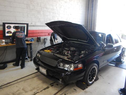 2003 Ford Crown Victoria - black Ford sedan on rollers in a dyno shop sits hood-up while an operator watches a monitor at left.