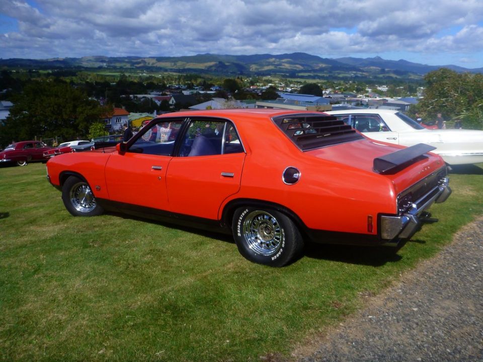 Orange sedan with louvers on the rear window and a spoiler parked on grass with other cars and a town in the background