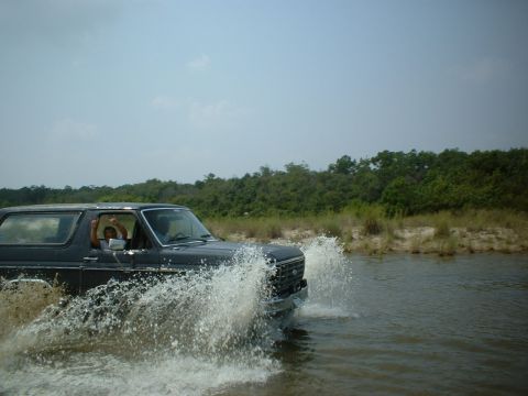 1985 Ford Bronco - A dark 1980s Ford Bronco navigates through shallow water, creating a large splash.