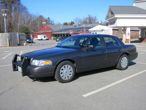 2001 Ford Crown Victoria - A dark gray 2001 Ford Crown Victoria police car is parked in a lot with buildings in the background.