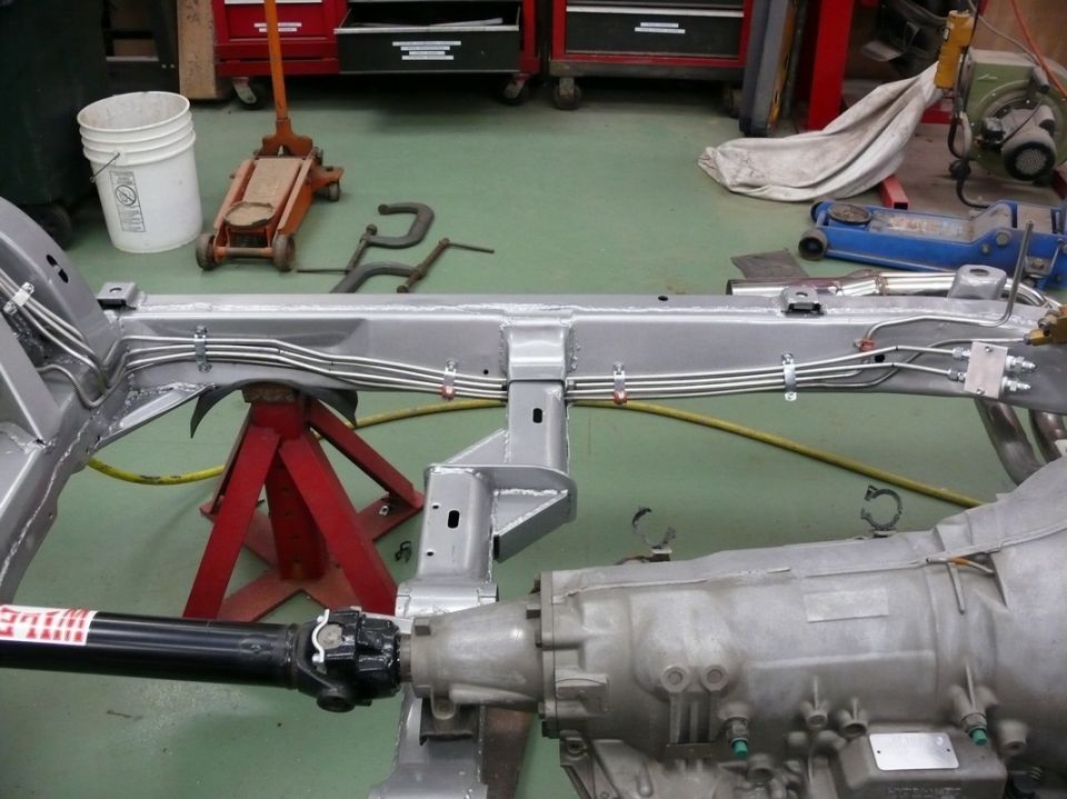 Car frame on jack stands with transmission and driveshaft visible. Some brake lines are routed along the frame