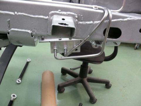 Vehicle frame with brake lines and suspension parts, on a green floor with a rolling chair