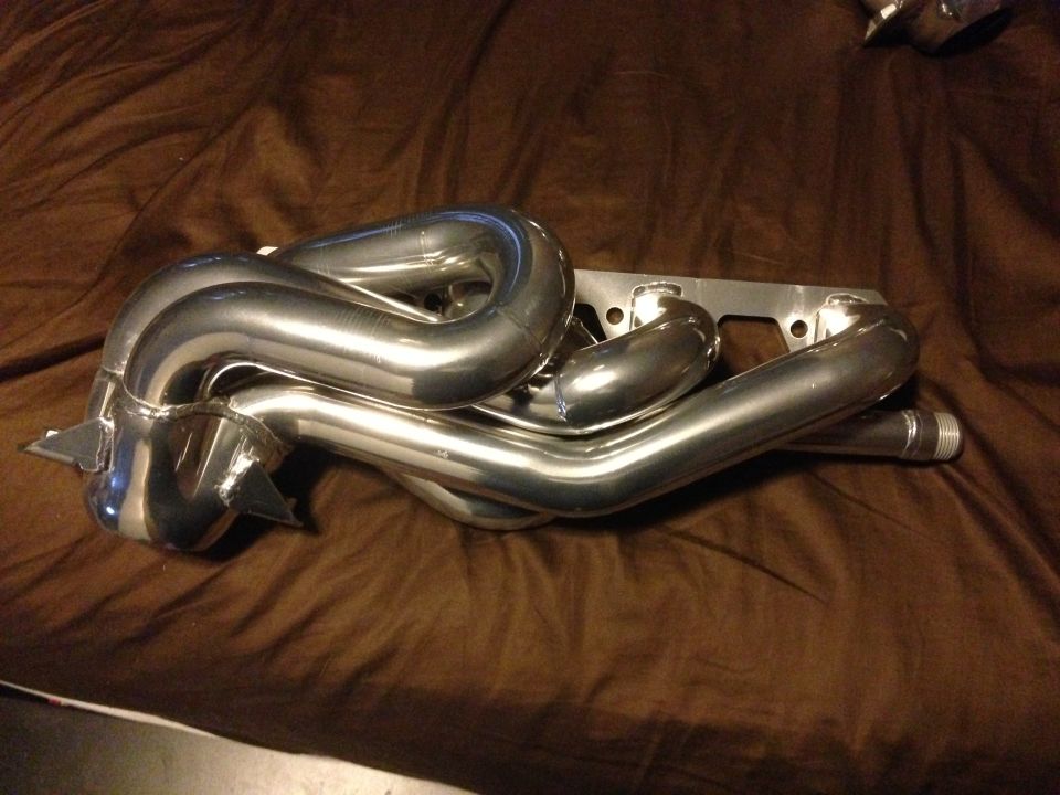 Shiny metal exhaust headers on a brown cloth surface. Text at bottom reads SUPERMOTORS