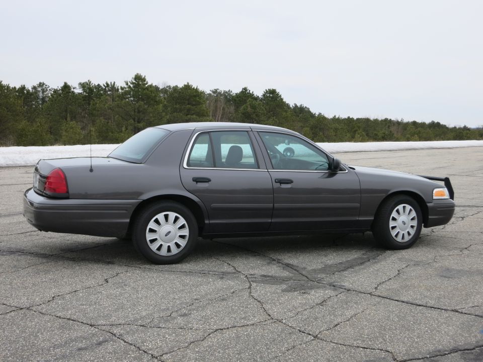 Side view of a dark grey Ford Crown Victoria parked on cracked asphalt with trees and snow in the background