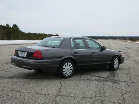 2001 Ford Crown Victoria - A dark grey 2001 Ford Crown Victoria sedan is parked on an empty asphalt lot with trees in the background.