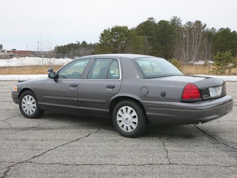 2001 Ford Crown Victoria - A grey 2001 Ford Crown Victoria is parked on cracked asphalt with trees in the background.