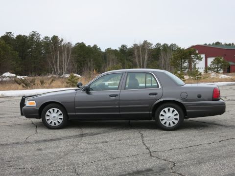 2001 Ford Crown Victoria - A dark grey 2001 Ford Crown Victoria Police Interceptor is parked on cracked asphalt with trees and a building in the background.