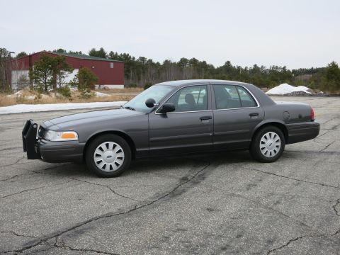 2001 Ford Crown Victoria - A dark grey Ford Crown Victoria sedan with a push bumper is parked on a cracked asphalt lot.