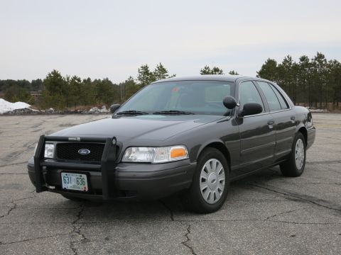 2001 Ford Crown Victoria - A dark grey 2001 Ford Crown Victoria, equipped with a push bar, is parked on a cracked asphalt surface.