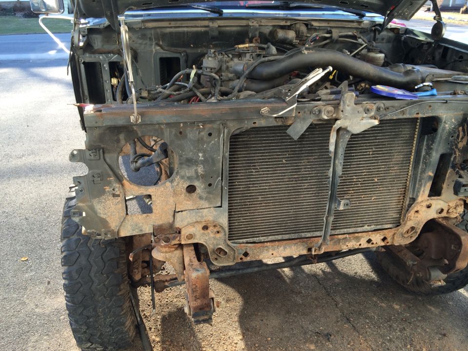 Front of truck with grille, bumper, and fender removed, exposing radiator, engine bay, and rusty frame during disassembly