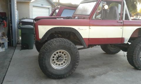 1968 Ford Bronco - A red and white Ford Bronco with a roll cage and oversized tires is parked in front of a garage.