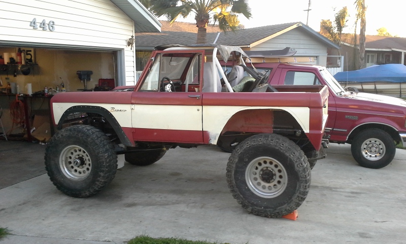 Red and white lifted Ford Bronco with large tires parked next to a red pickup truck in a driveway