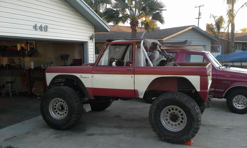 Red and white Ford Bronco with lifted suspension and large tires parked in front of a garage