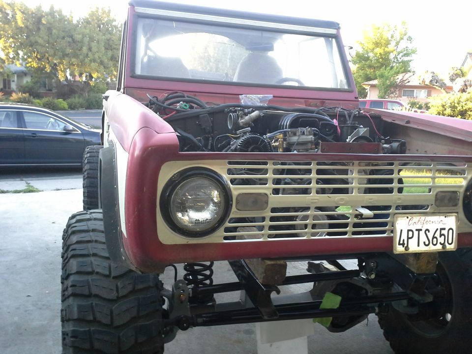 Front view of a red 1968 Ford Bronco with its hood open, showing the engine. It has large off-road tires and a California license plate reading 4PTS650