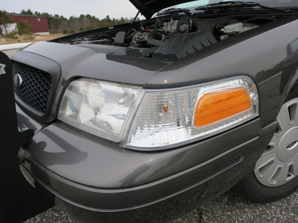 Front corner of a grey Ford Crown Victoria with hood open, showing engine. Headlight and orange turn signal visible