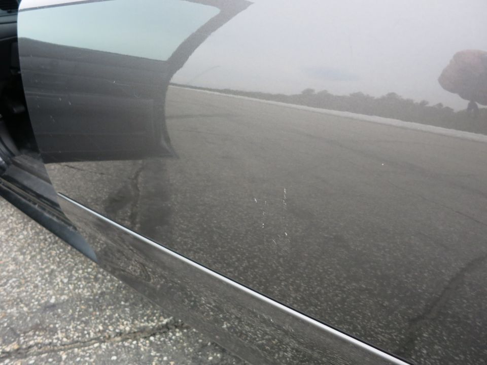 Gray car door with scratches and reflections of a road and trees