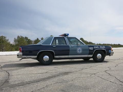1991 Ford Crown Victoria - A Massachusetts State Police car is parked on a cracked asphalt surface with trees and a cloudy sky in the background.