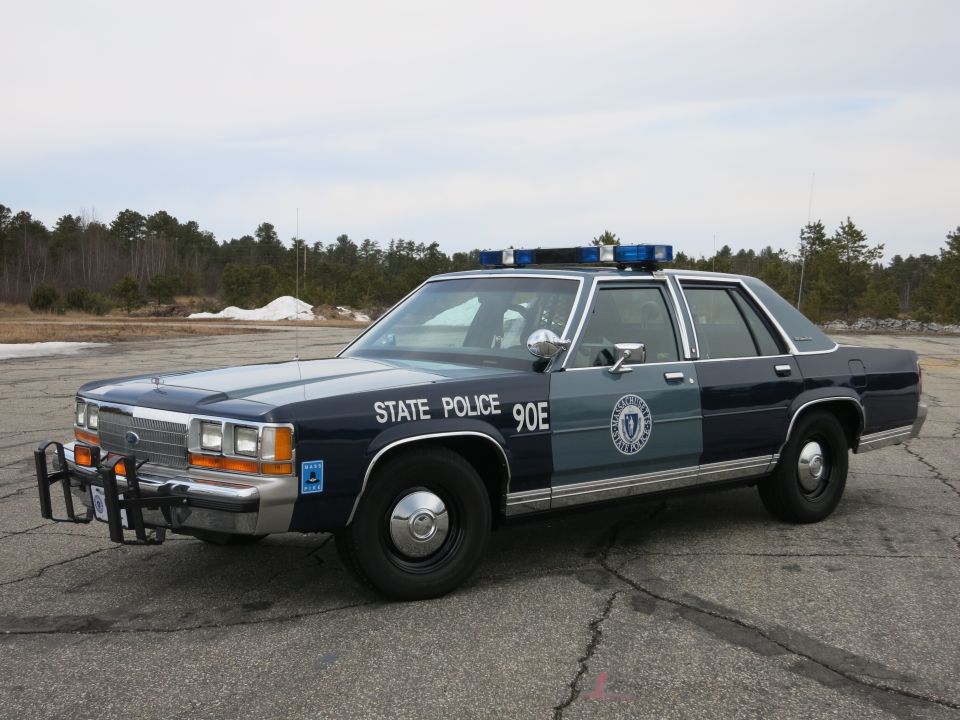 Front three-quarter of a blue-and-gray State Police Ford Crown Victoria sedan with push bar, marked 90E