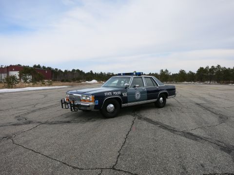 1991 Ford Crown Victoria - This 1991 Ford Crown Victoria police car is parked on an asphalt lot with trees and buildings in the background.