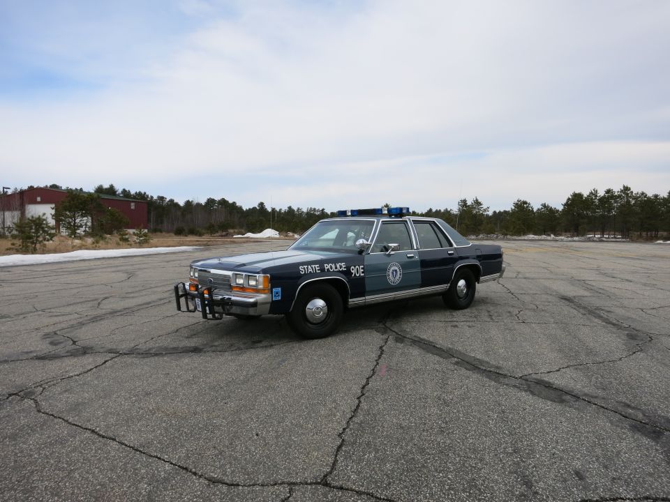 Distant front three-quarter of a blue-and-gray State Police Ford sedan on open cracked pavement