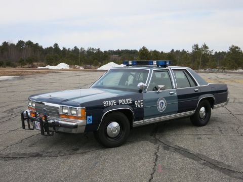 1991 Ford Crown Victoria - A dark blue and gray Ford Crown Victoria police car is parked on an asphalt lot with trees and snow in the background.