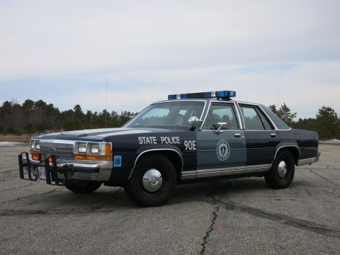 1991 Ford Crown Victoria - A 1991 Ford Crown Victoria police car, marked "STATE POLICE 90E", is parked on an outdoor lot under a cloudy sky.