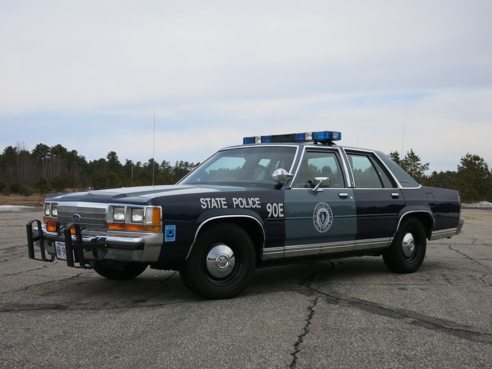 Front three-quarter of a blue-and-gray State Police Ford sedan marked STATE POLICE 90E on pavement
