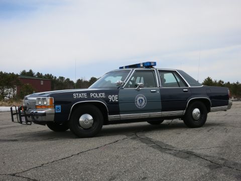 1991 Ford LTD Crown Victoria - Driver-side front three-quarter view of a navy-and-gray Massachusetts State Police Ford cruiser with push bar and STATE POLICE 90E letterin…