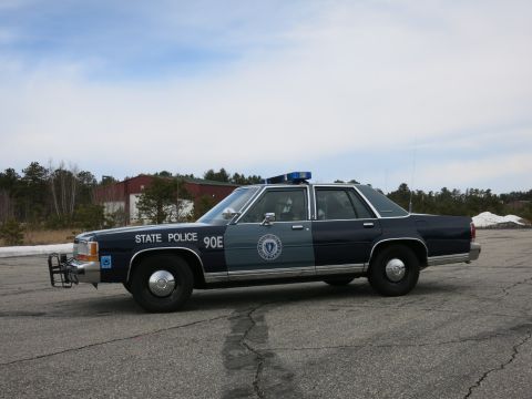 1991 Ford LTD Crown Victoria - Full driver-side profile of a two-tone navy and gray Massachusetts State Police Ford cruiser parked on cracked pavement under a blue sky.