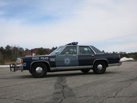 1991 Ford LTD Crown Victoria - Driver-side profile of a navy-and-gray Massachusetts State Police Ford cruiser with roof light bar and STATE POLICE 90E door markings.