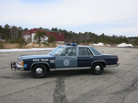 1991 Ford LTD Crown Victoria - two-tone Massachusetts State Police Ford cruiser sits front-three-quarter on a paved lot with a red warehouse and pine trees behind.
